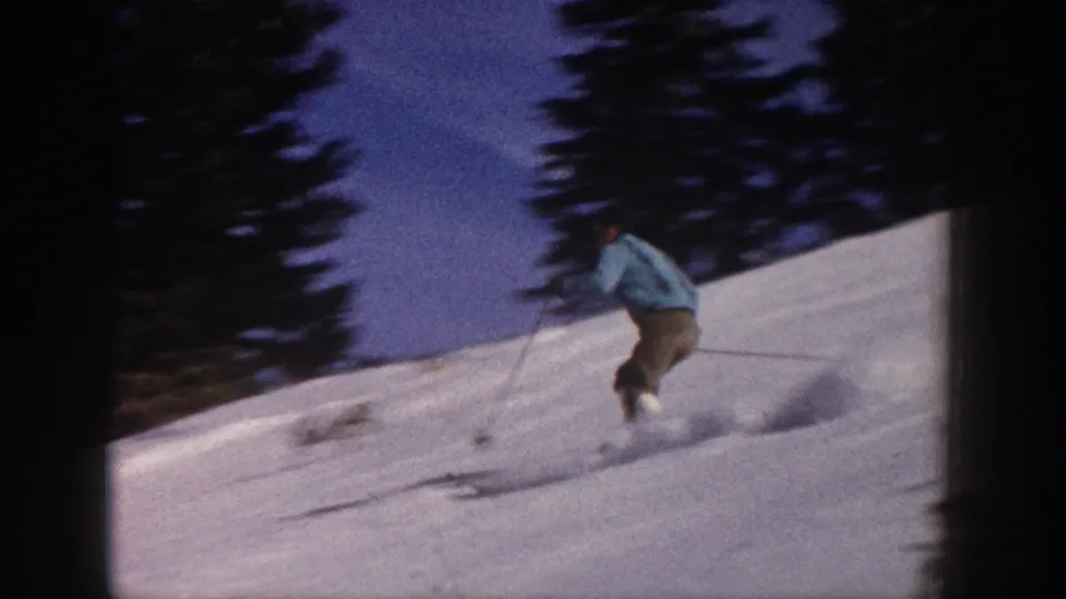 An adult skier in a light blue jacket and brown pants turns on an Aspen snow slope, with evergreen trees behind him in faded 8mm color.