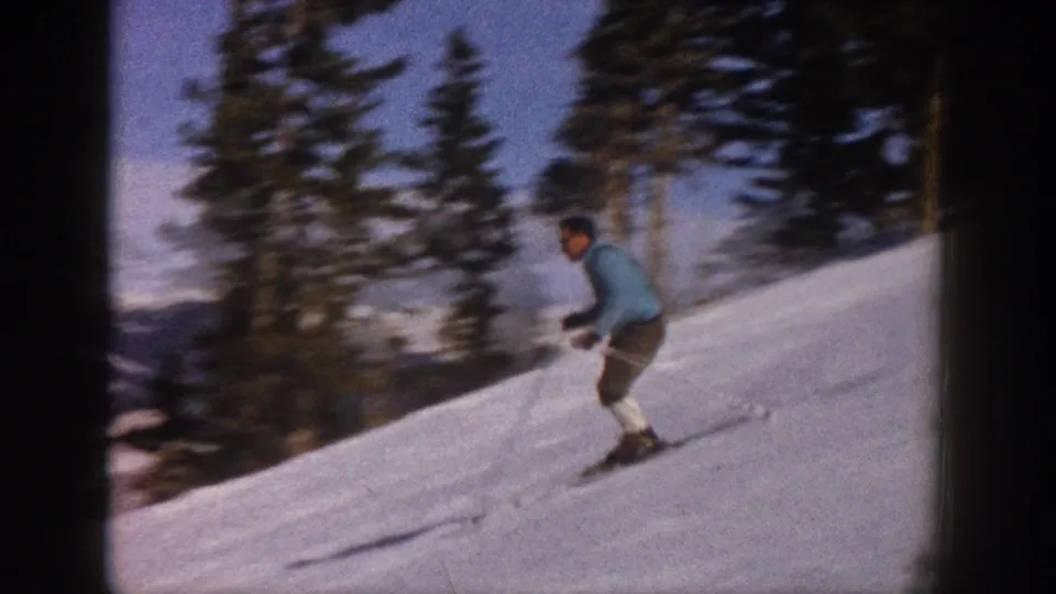 The skier continues downhill on an Aspen snow slope, passing evergreen trees in a soft, slightly blurred 1961 color 8mm frame.