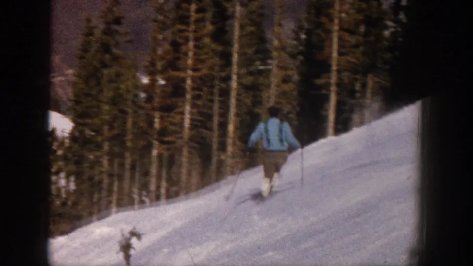 1961 Aspen 8mm frame with the skier seen from behind on a white slope bordered by tall evergreen forest.