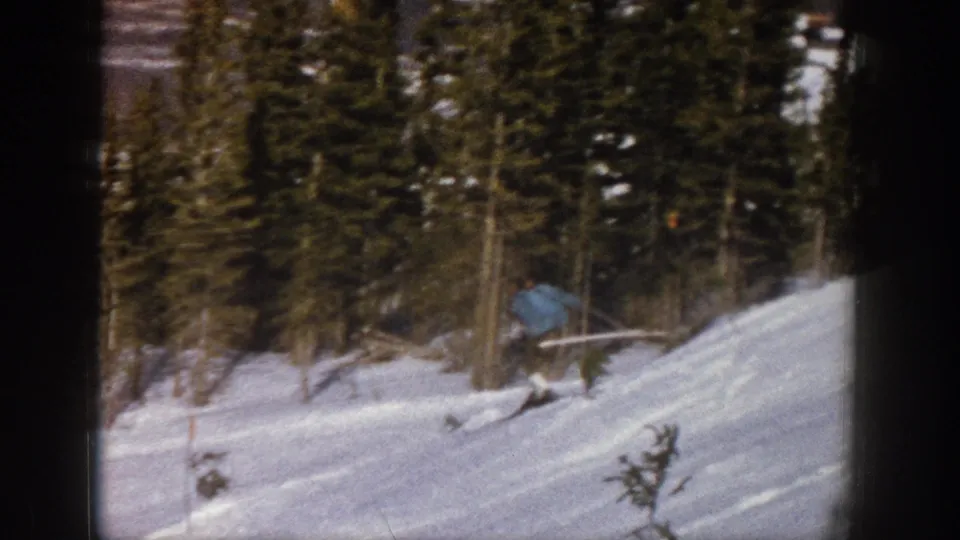 1961 Aspen 8mm frame showing the skier moving across a snowy run with small saplings and dark evergreen forest behind.