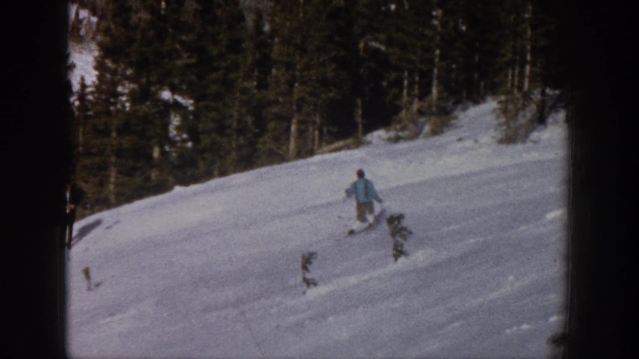 A skier continues across a snowy hillside with trees in the background.