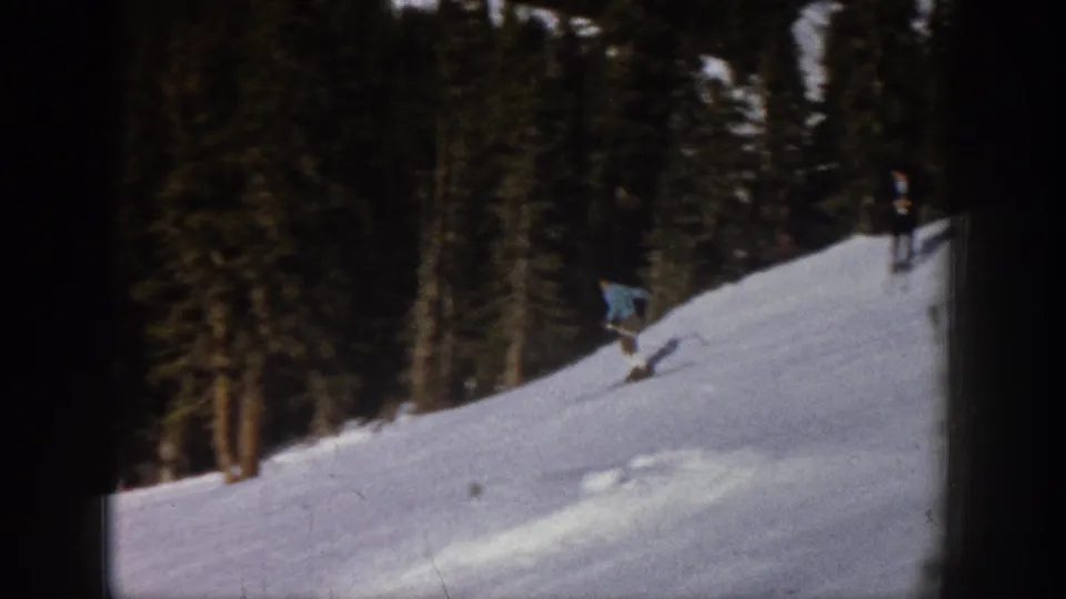 The skier continues downhill beside tall evergreens, with another distant person visible higher on the snowy Aspen slope.