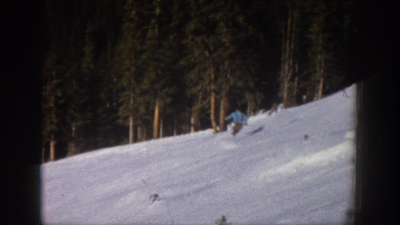 1961 Aspen home-movie frame showing a distant skier moving across a snow-covered hillside bordered by tall evergreens.