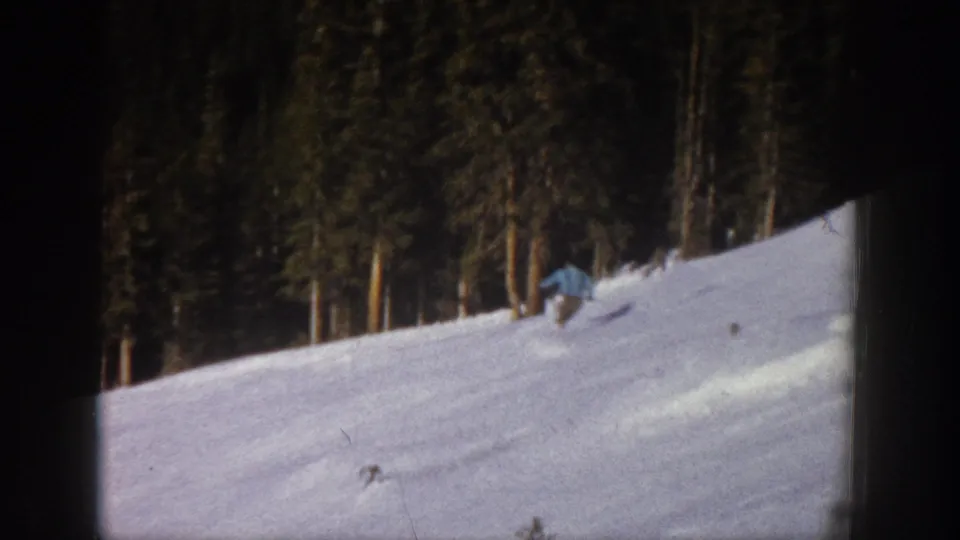 1961 Aspen home-movie frame showing a distant skier moving across a snow-covered hillside bordered by tall evergreens.