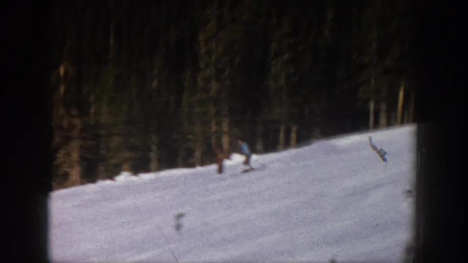 Home-movie view of a skier in pale clothing crossing an Aspen hillside, with evergreen trees and 8mm film edges visible.