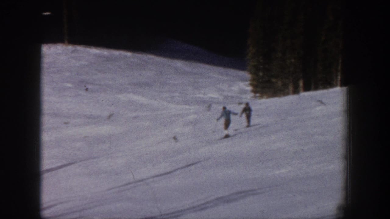 Two skiers move downhill on a snowy Aspen slope in 1961 home-movie footage, with trees at the upper edge of the run.