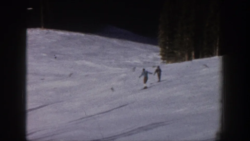 Two skiers move downhill on a snowy Aspen slope in 1961 home-movie footage, with trees at the upper edge of the run.