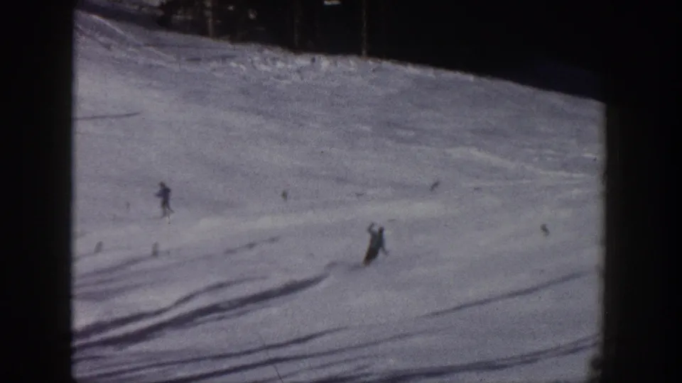 Distant skiers traverse a sunlit snow slope in Aspen, Colorado, with tracks and shadows visible across the run.