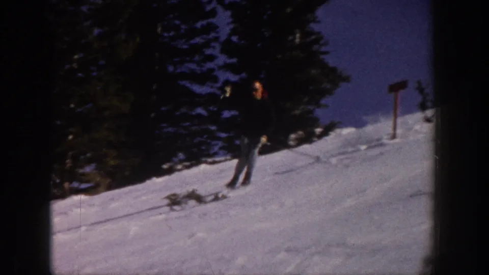 A skier in light pants appears near evergreen trees and a slope marker on a snowy Aspen hillside in 1961 home-movie footage.