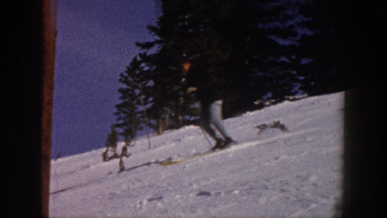 1961 Aspen home-movie frame showing a skier in a dark jacket and light pants moving across a snowy slope near evergreens.