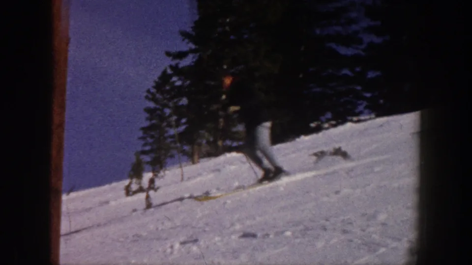 1961 Aspen home-movie frame showing a skier in a dark jacket and light pants moving across a snowy slope near evergreens.