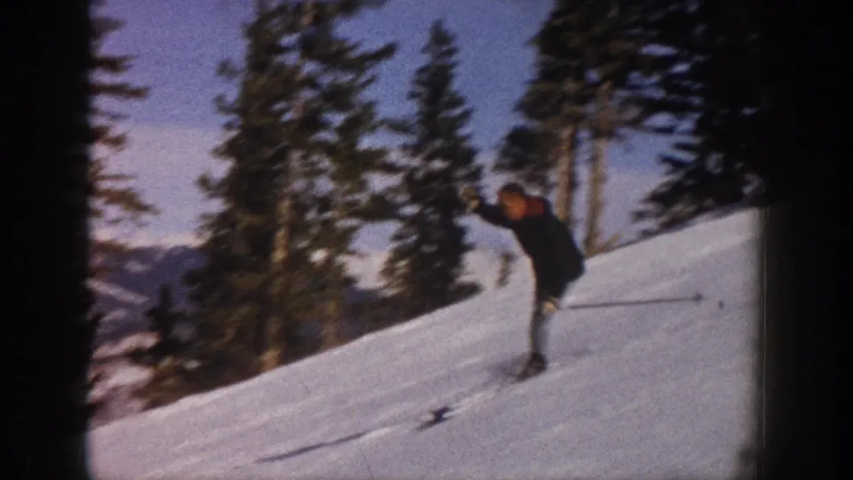 1961 Aspen home-movie frame of a skier descending a sunny slope, with evergreen trees and distant mountain terrain visible.