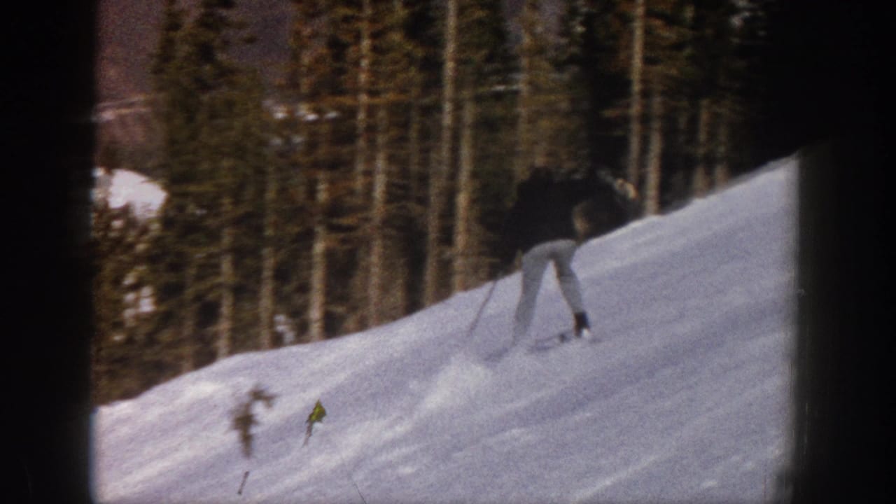 In the supplied 1961 Aspen context, a skier in a dark jacket and light pants moves across a snowy slope bordered by evergreens.