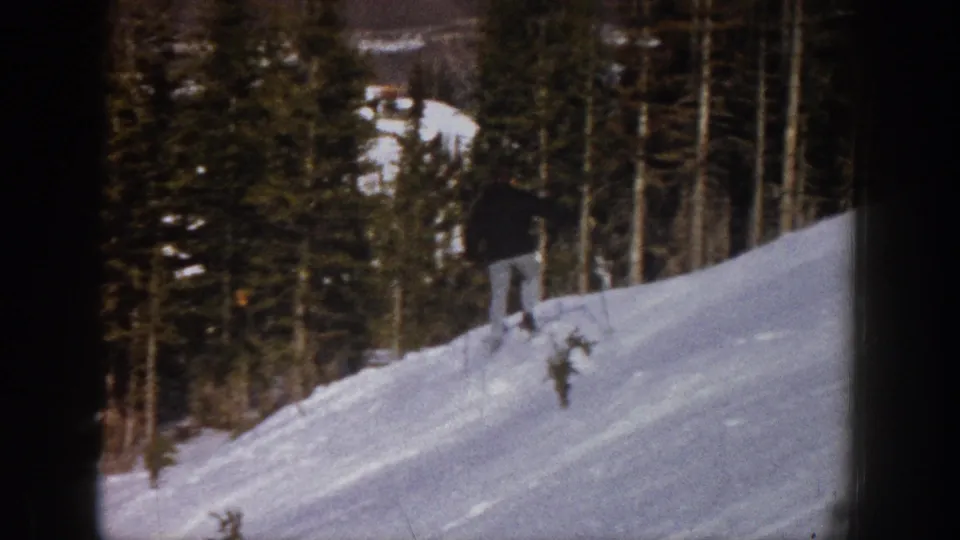 The skier moves farther across the Aspen snow slope, framed at a distance against dense evergreen trees.