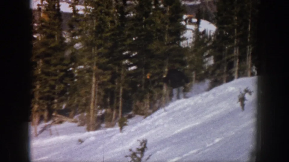 A distant skier approaches the wooded edge of the slope in the 1961 Aspen home-movie footage, with motion blur and 8mm grain visible.