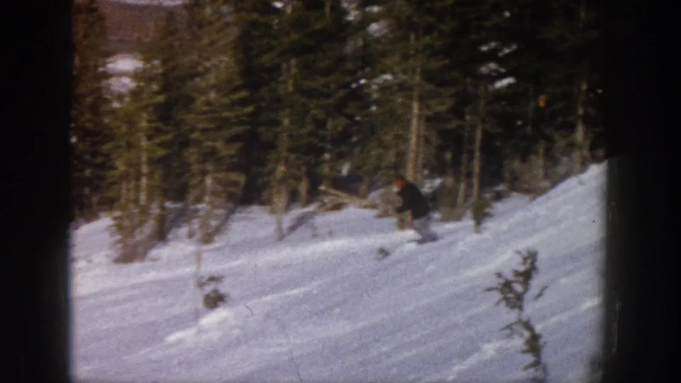 The skier leans into the descent on a snowy Aspen hillside, with small saplings and evergreen forest visible in the background.