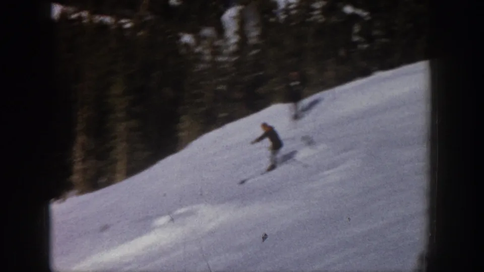 The skier continues across a bright snowy hillside, with dense evergreen trees lining the upper slope.