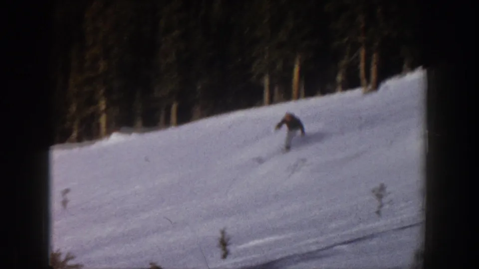 A small skier figure descends a broad snow-covered slope, partly softened by the vintage 8mm film image.