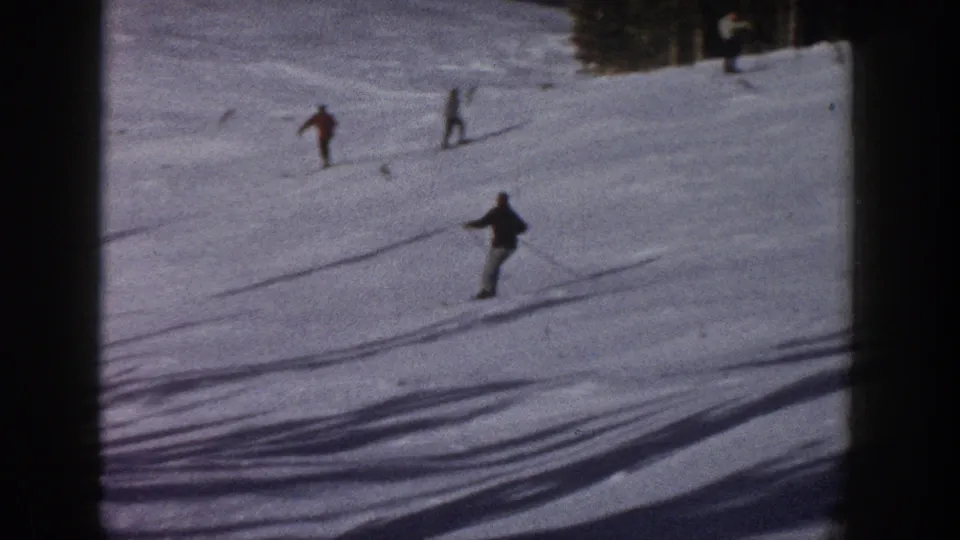 1961 Aspen home-movie frame showing distant skiers moving across a snowy slope bordered by dark evergreens.