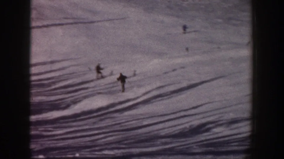 1961 Aspen 8mm frame with several distant skiers spread across a broad snowfield and dark shadows.
