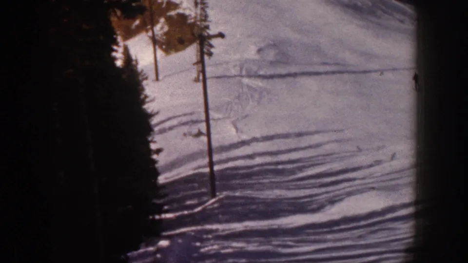 1961 Aspen ski-slope frame with a pole at the run edge, dark pines, long shadows, and distant activity on the snow.
