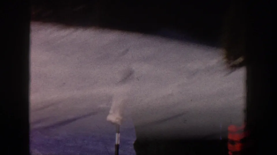 Soft, blurred 8mm frame of the Aspen ski slope, with snow, shadows, and a small foreground marker visible.