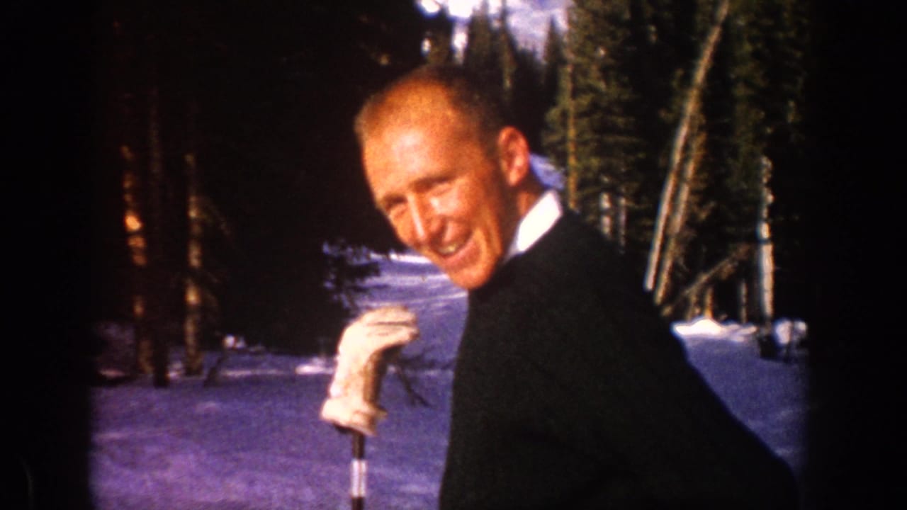 Smiling man in a dark suit stands in snowy woods with a coat over his shoulder.