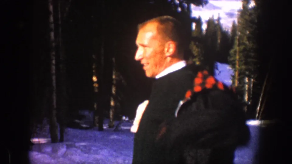 Side view of the man putting on a plaid-lined coat in the snowy Aspen forest, with dark trees behind him.