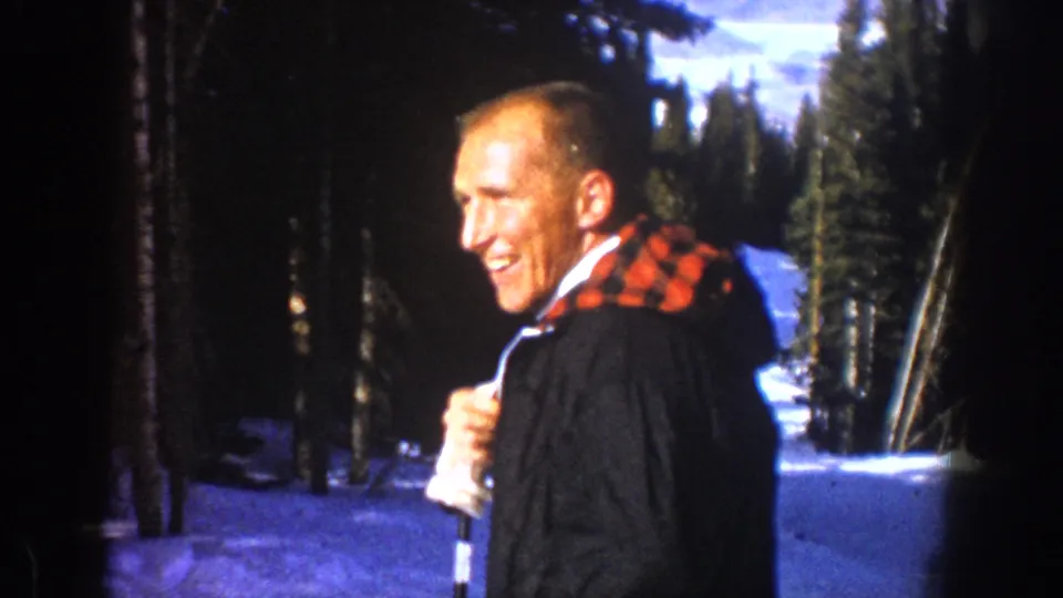 The man smiles toward the camera in 1961 Aspen, holding a walking stick with snowy evergreens behind him.