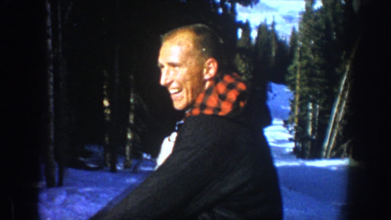 In 1961 Aspen footage, a smiling male skier in a dark jacket and plaid fabric faces the camera near pine trees on snow.