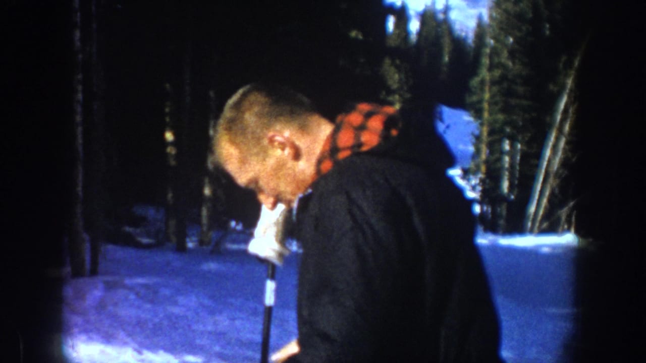 The skier in the Aspen home movie leans forward beside a ski pole, with snow and dark pine trees behind him.
