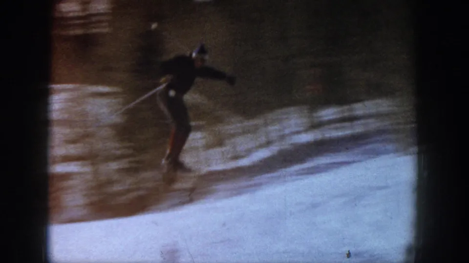 The skier appears mid-jump or crossing over a small ditch in the 1961 Aspen ski footage, with snow and tree shadows around him.