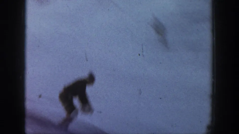 A distant skier in dark clothing bends forward on snow in faded 1961 Aspen 8mm home-movie footage.