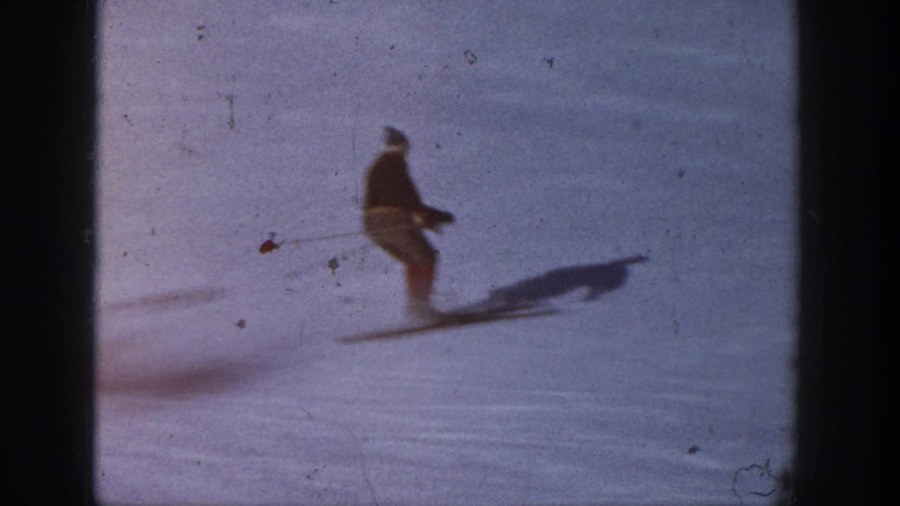 Skier in dark top and light pants gliding across snow with poles and a long shadow.