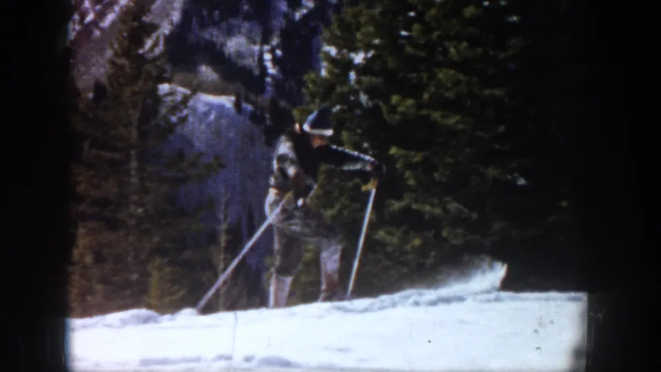 1961 Aspen home-movie frame showing a skier in a dark jacket and knit hat using poles on a snowy slope near evergreen trees.