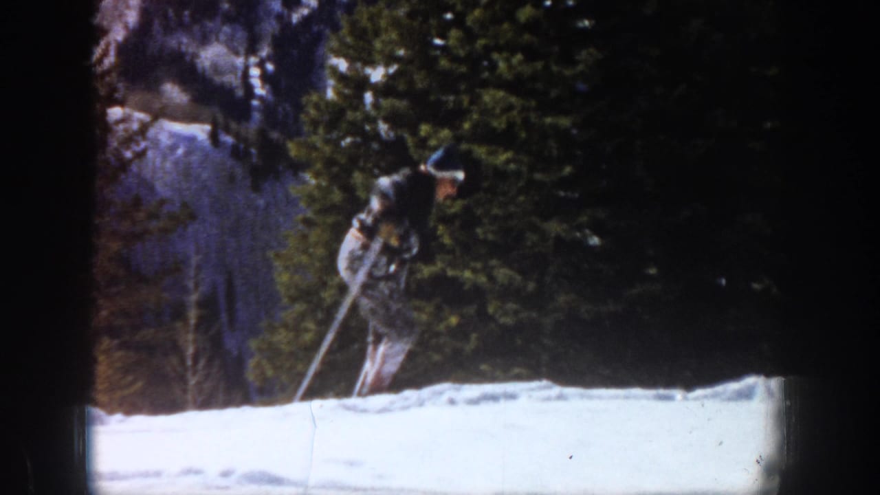 1961 Aspen frame with a lone skier using poles on bright snow, partly framed by evergreen trees and distant mountain terrain.