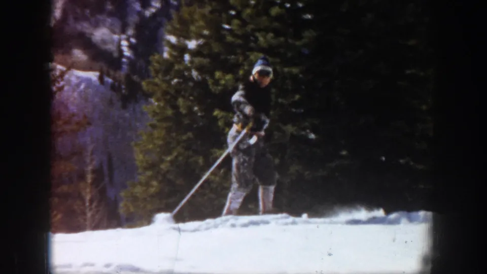 1961 Aspen home-movie frame showing the skier turned toward the camera on snow, with trees and a shaded mountain background.