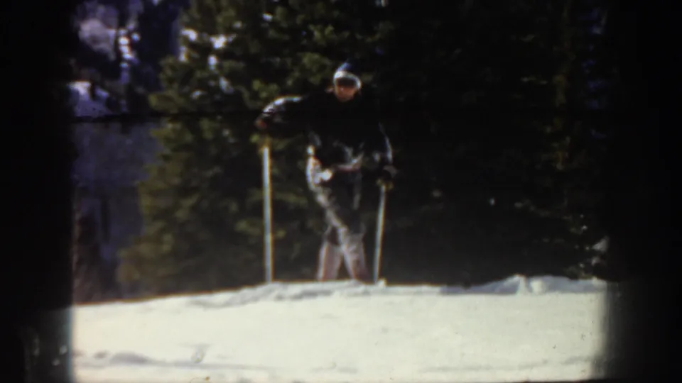 1961 Aspen frame showing the skier upright with poles on a snowy rise, backed by dark evergreen trees and heavy film shadowing.