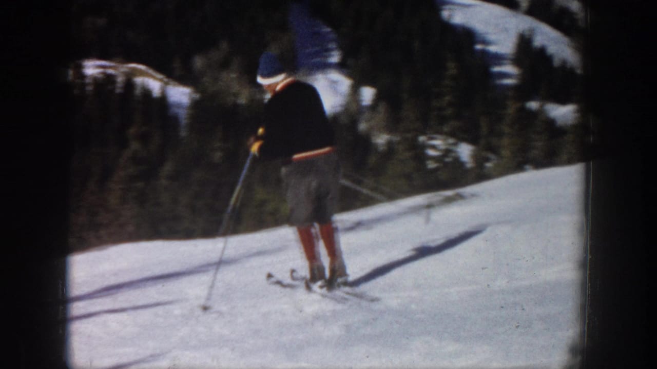 1961 Aspen home-movie frame showing a male skier paused on a bright snowy slope with ski poles and forest behind him.