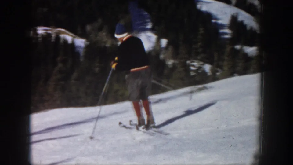 1961 Aspen home-movie frame showing a male skier paused on a bright snowy slope with ski poles and forest behind him.