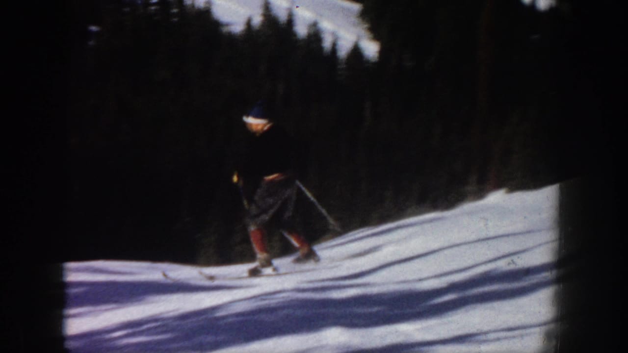 1961 Aspen footage frame showing a male skier descending a snowy hill with poles, dark trees, and bright snow shadows.