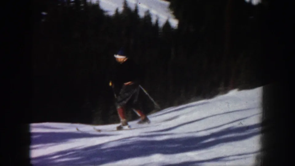 1961 Aspen footage frame showing a male skier descending a snowy hill with poles, dark trees, and bright snow shadows.