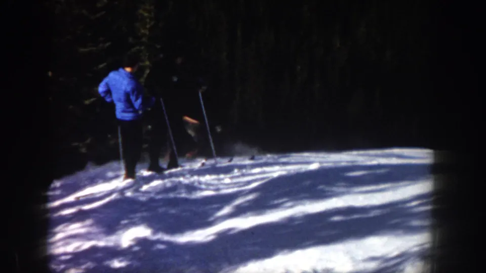1961 Aspen home-movie frame with a skier in a blue top viewed from behind on a shaded snowy slope near evergreens.