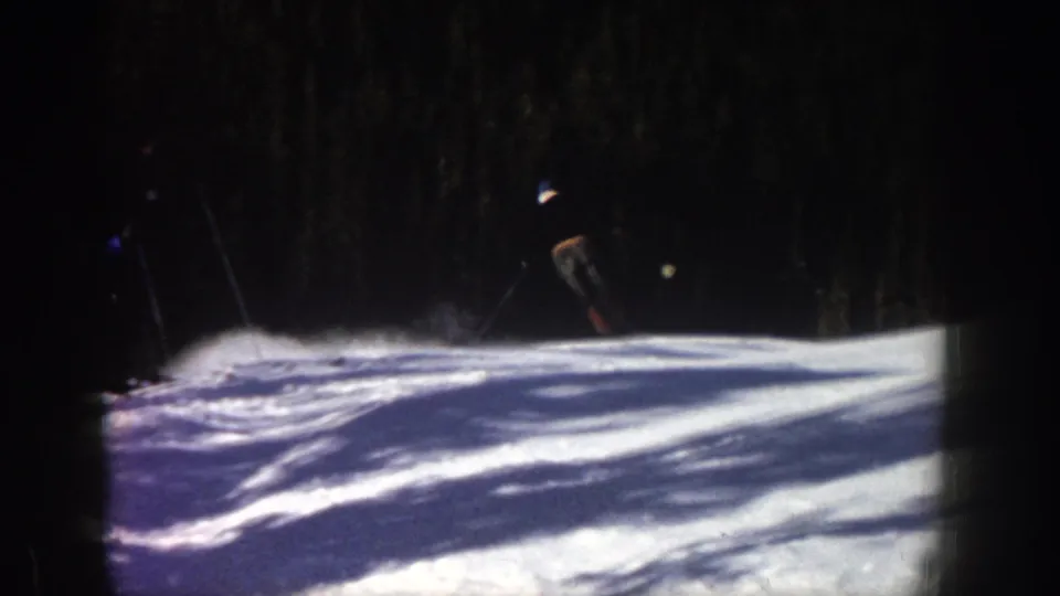 1961 Aspen frame showing a skier completing a turn on a snowy hill, with kicked-up snow and dense evergreens in the background.