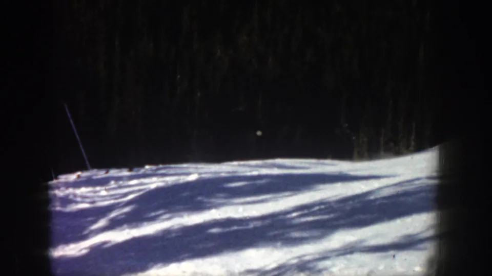 1961 Aspen home-movie frame showing a sunlit snowy slope below a dark evergreen forest, with no skier clearly visible in this moment.