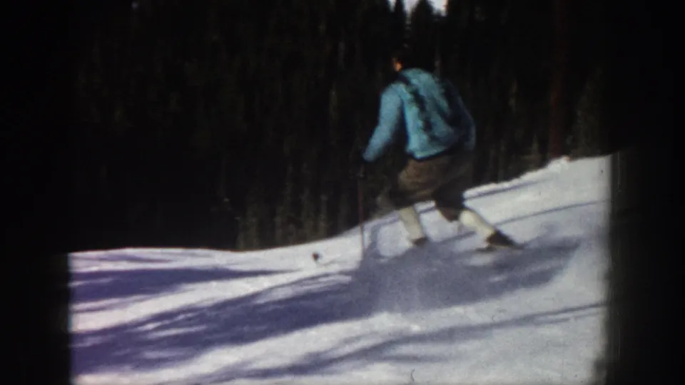 1961 Aspen home-movie frame showing a lone skier in a light blue jacket and brown pants using poles on a sunny snowy slope.