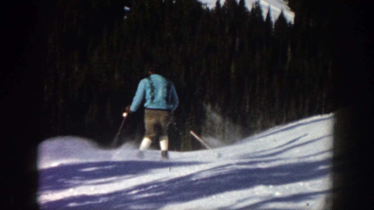 In Aspen, Colorado, 1961, an adult skier in a light sweater, shorts, suspenders, and high socks moves downhill on a bright snowy slope.