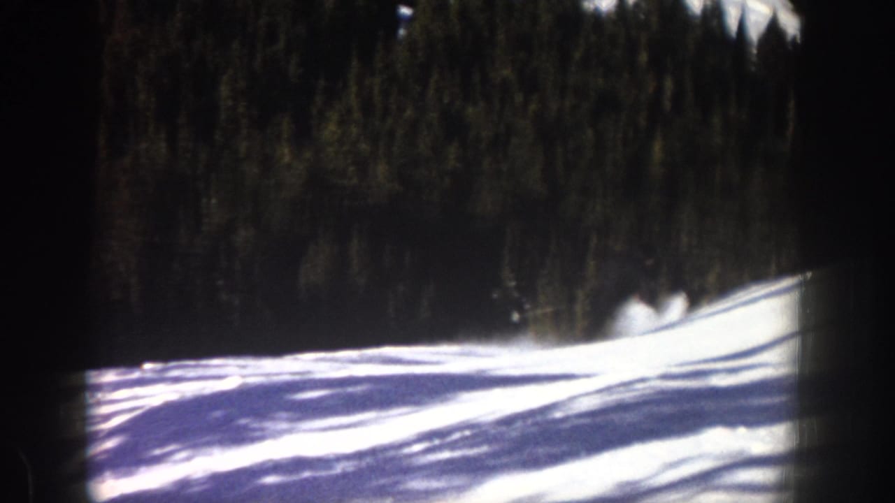1961 Aspen ski footage frame with snow, forest backdrop, strong shadows, and possible skier movement near the right side.