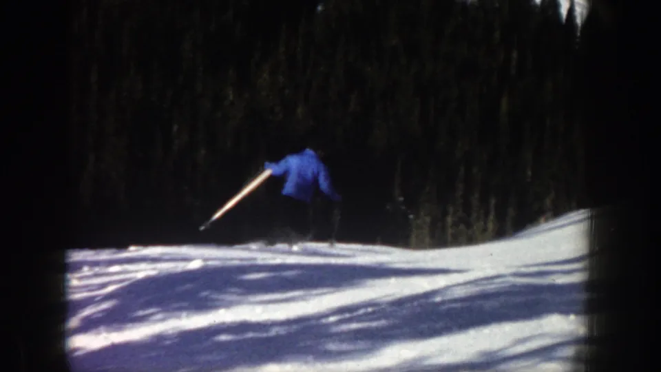 1961 Aspen home-movie frame of a distant skier in a blue jacket moving downhill across a snowy slope near evergreens.
