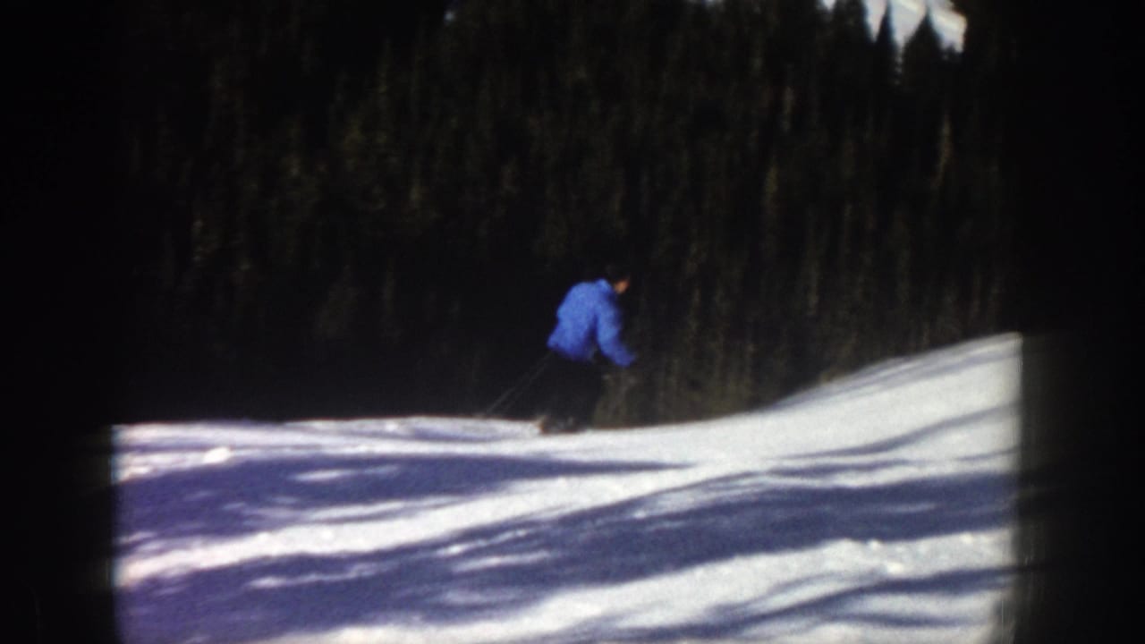 1961 Aspen home-movie frame showing a skier in a blue jacket moving across a snow-covered slope with evergreen forest behind.